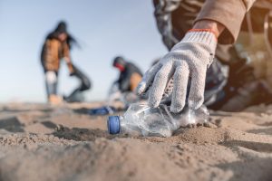 a hand reaching down to pick up a plastic water bottle on the beach, two other people are in the distance also cleaning the beach.