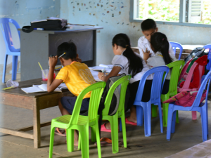 four children sitting in a classroom on colourful plastic chairs. each have their back to the camera and are looking down at papers on the table in front of them.