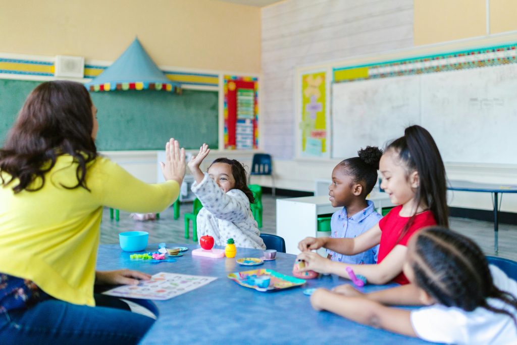 four young kids playing with a teacher in a classroom. The image is used to represent the benefits of investing in education