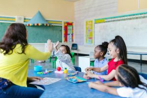 four young kids playing with a teacher in a classroom. The image is used to represent the benefits of investing in education