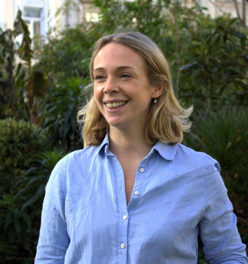 headshot of cate quentin naturally looking off to the side. she is outside with greenery visible in the background.