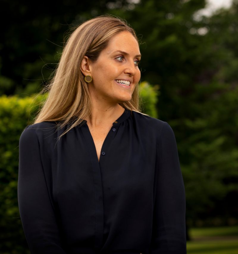 headshot of louise blanc naturally looking off to the side. she is outside with greenery visible in the background.