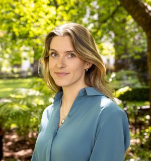 Professional headshot of Caitlin taken in a park with greenery visible behind her