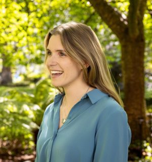 relaxed, natural headshot of Caitlin taken in a park with greenery visible behind her. She is smiling looking off to the left.